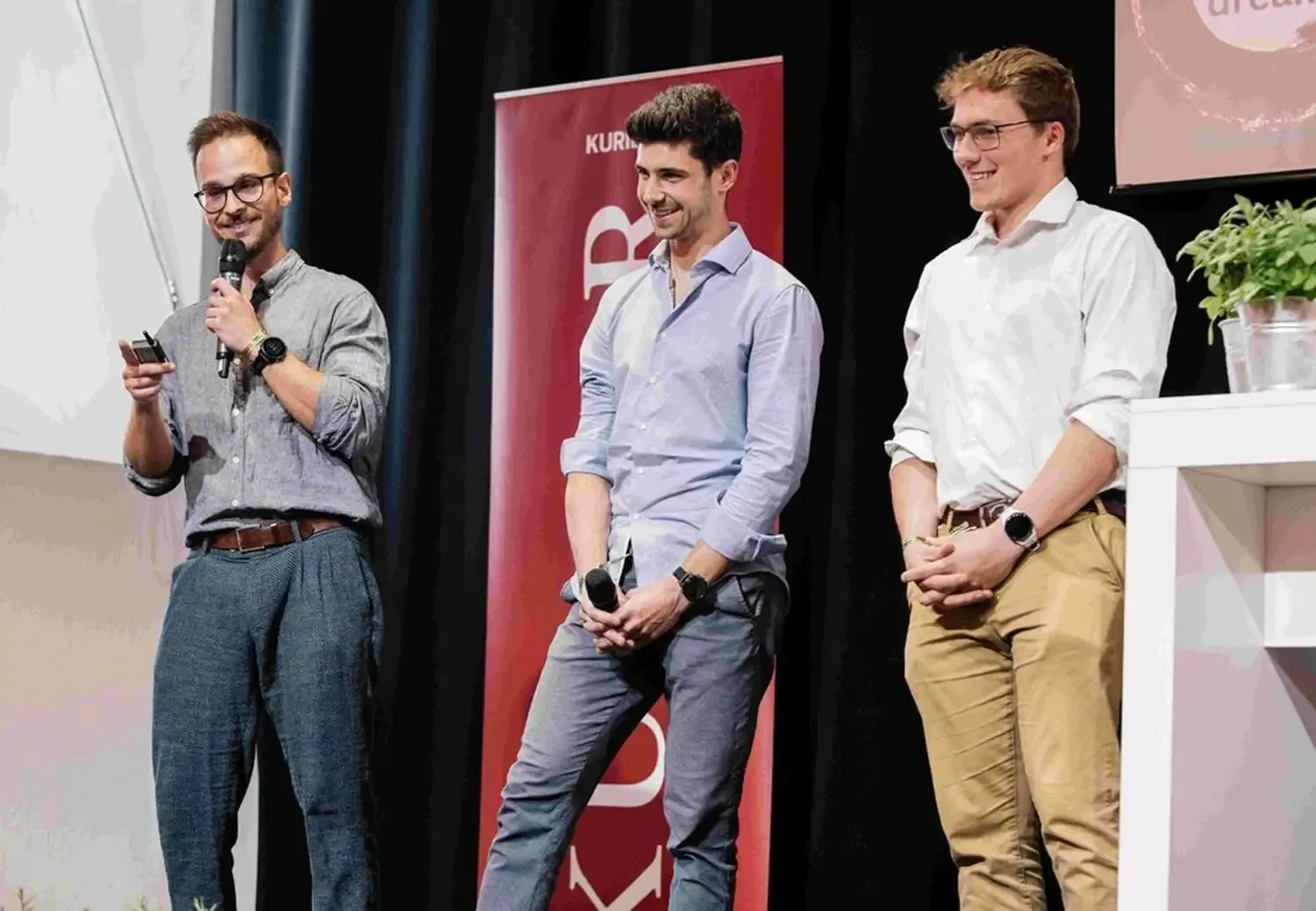 Drei Männer stehen lächelnd auf einer Bühne, einer hält ein Mikrofon, im Hintergrund ein roter Kurier-Banner
