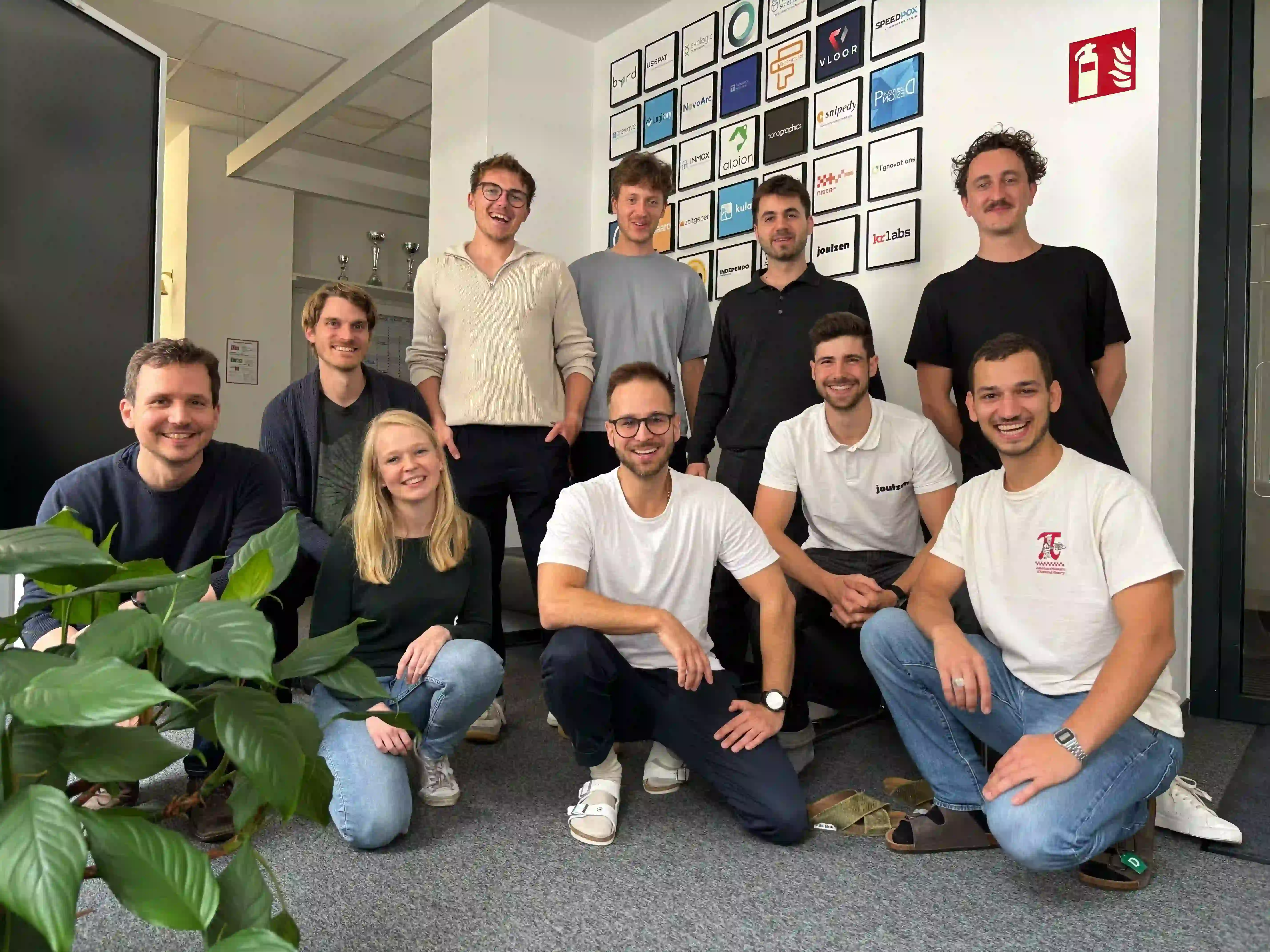 Zehn Teammitglieder posieren im Büro, einige stehend, andere kniend, vor einer Wand mit Startup-Logos