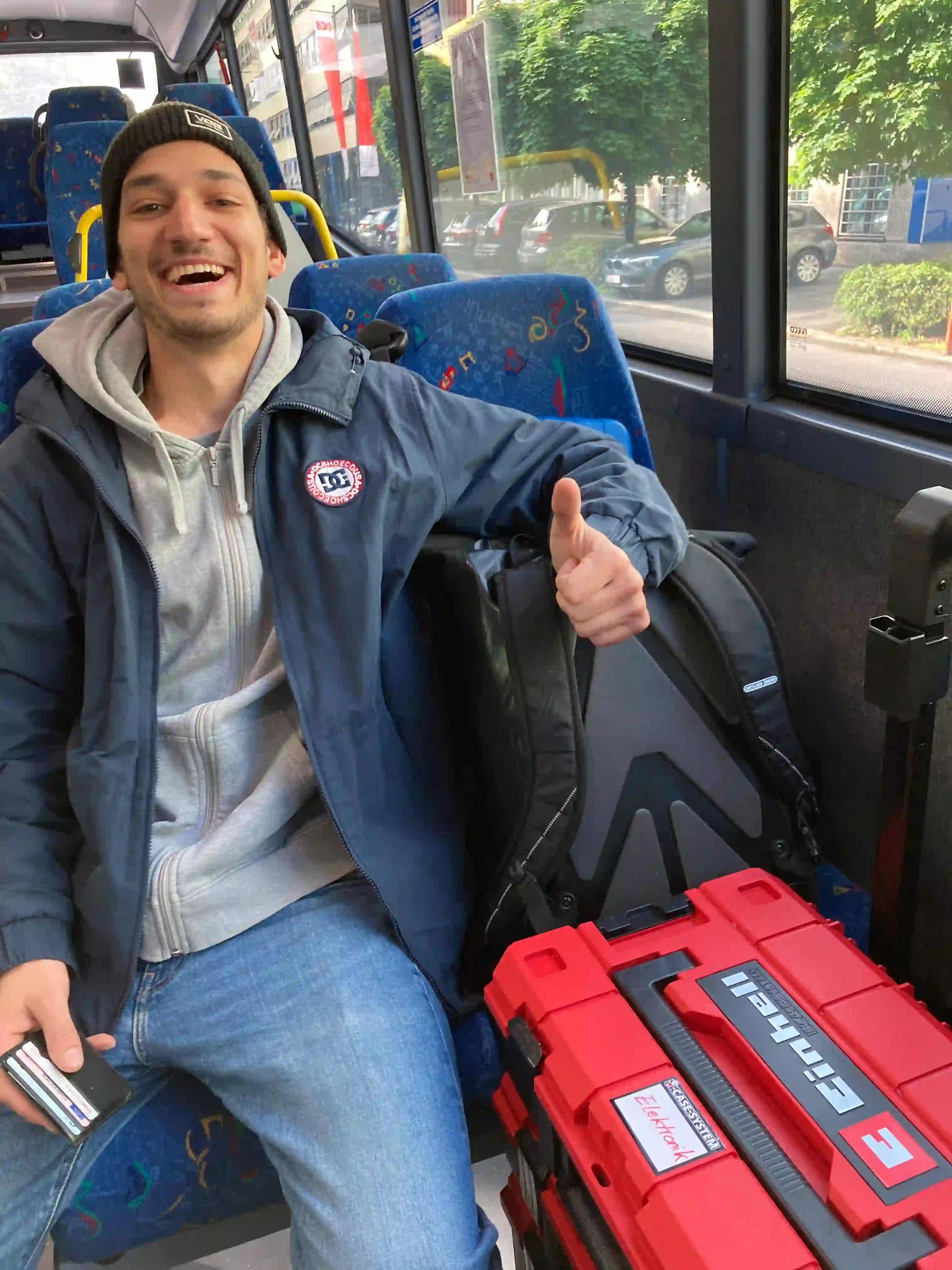 Junger Mann mit Mütze sitzt lachend im Bus, zeigt Daumen hoch, neben ihm rote Werkzeugkoffer