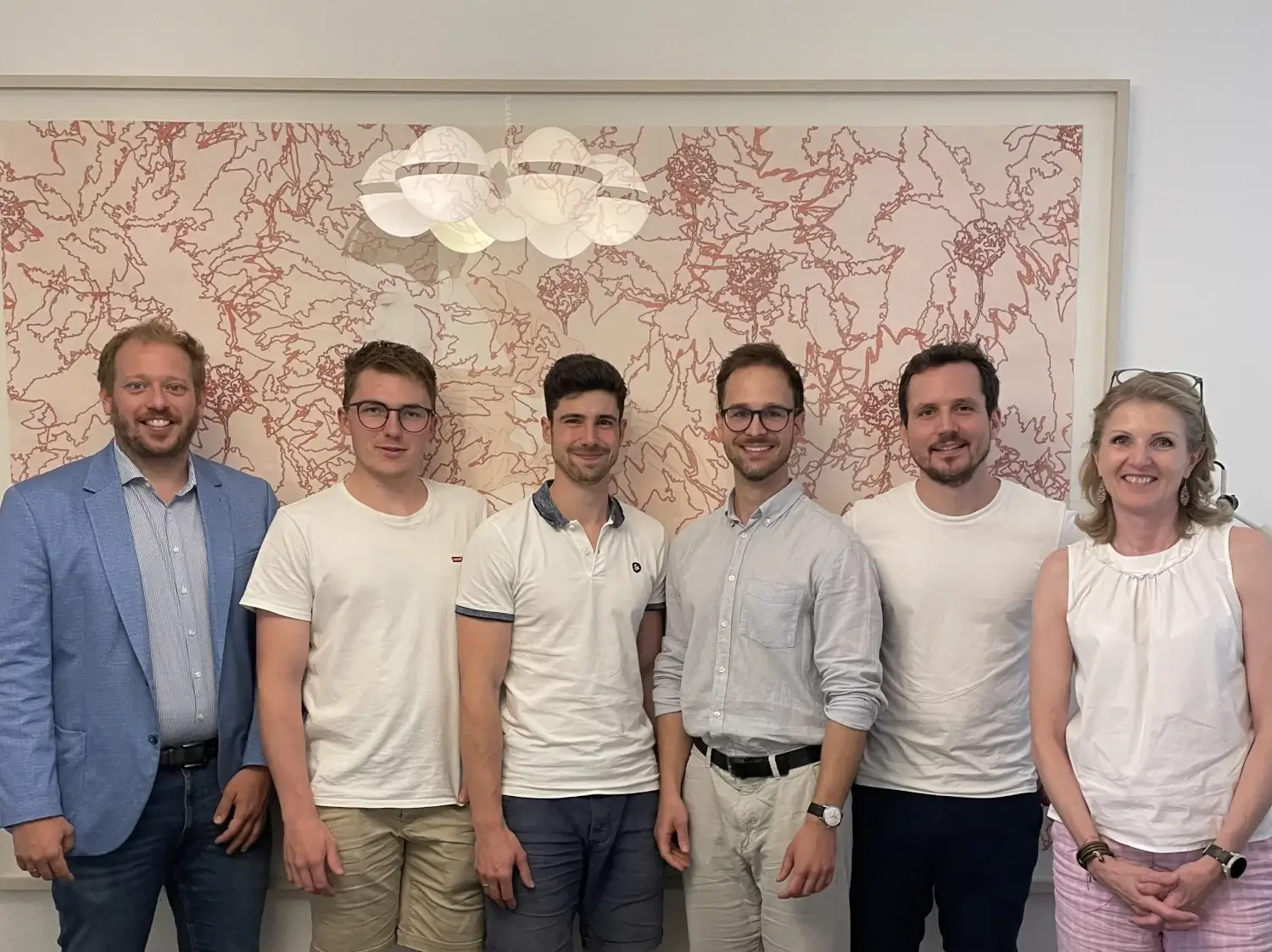 Sechs Personen stehen nebeneinander vor einem dekorativen Kunstwerk an der Wand